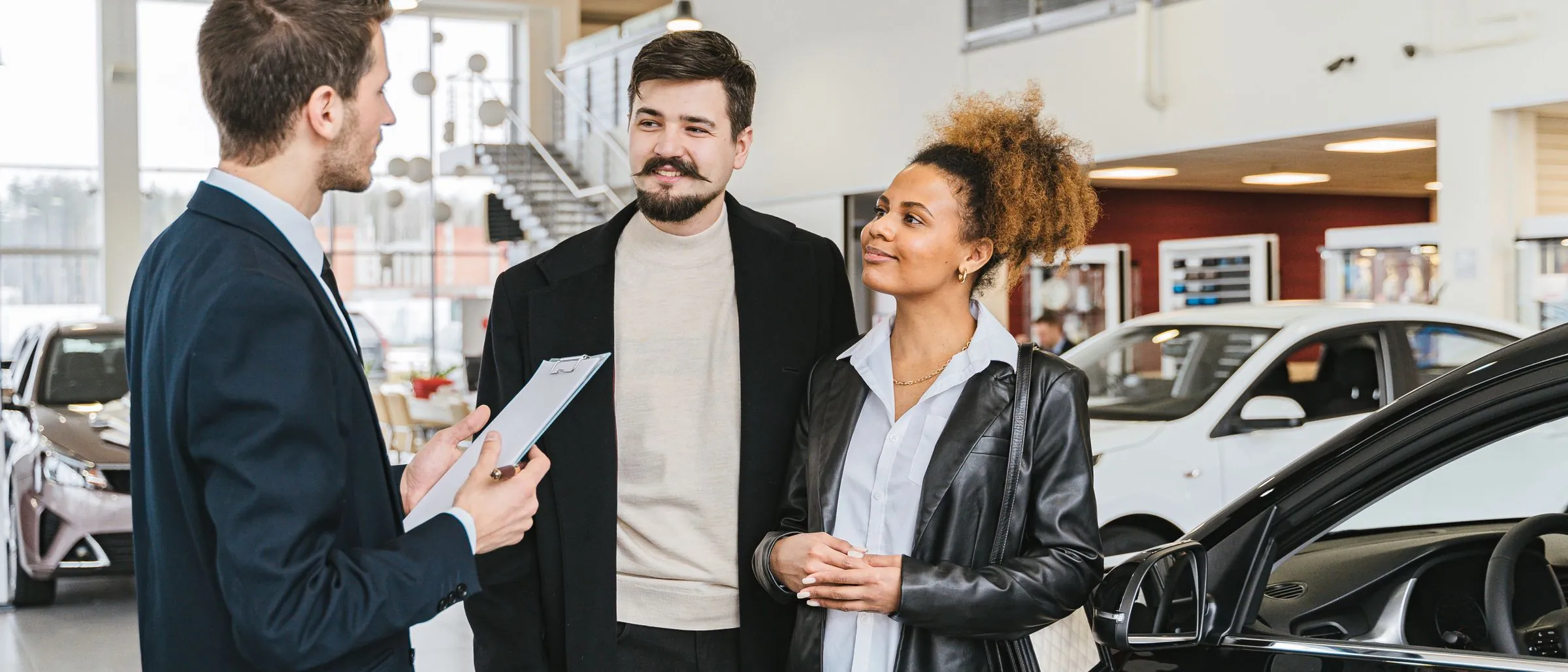 A salesman with a clipboard talking to a couple in a car showroom.