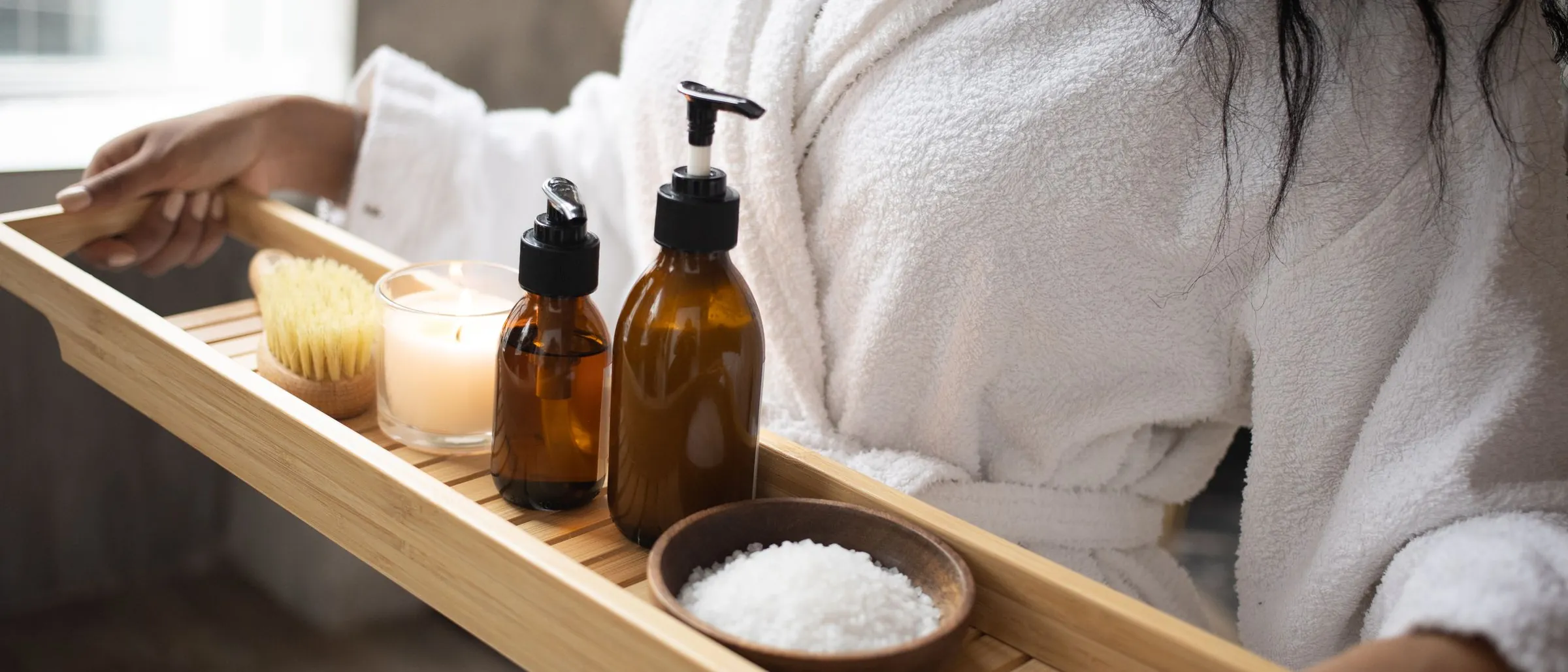A woman in a white robe carrying a bath tray with amber skincare bottles, a candle and bath salts.
