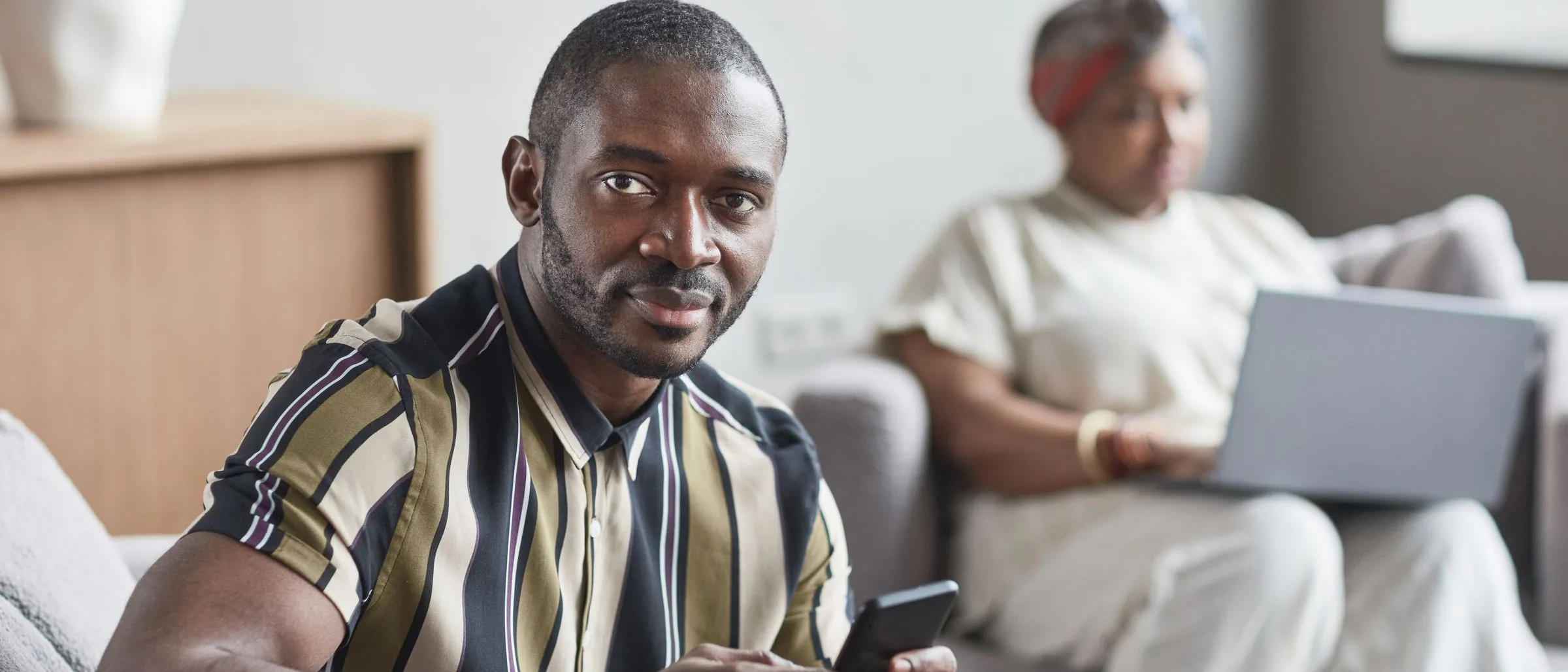 A man holding his phone at home, with a woman working on a laptop behind him.